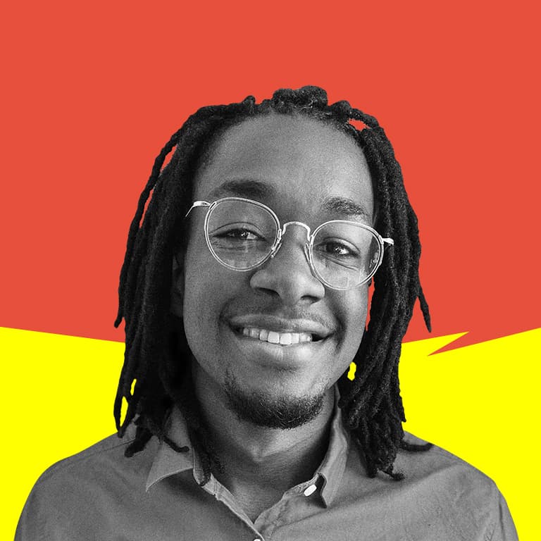 A young, African-American male with long black dreads and wearing a pink graphic t-shirt and glasses, smiling into the camera.