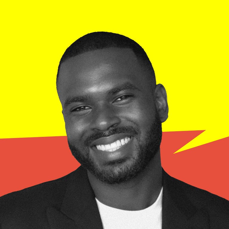 A young Black American man with a haircut known as the low-cut Caesar wearing a white t-shirt and double-breasted black blazer smiling